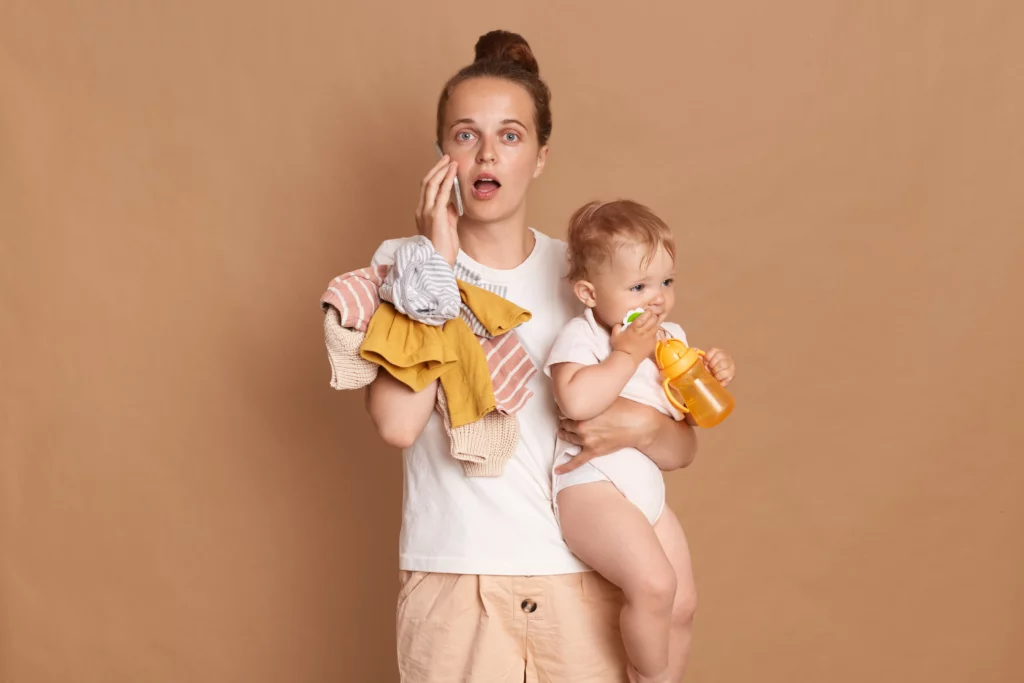 Portrait d'une belle femme choquée avec une coiffure en chignon portant un t-shirt blanc debout avec sa petite fille et parlant au téléphone, posant la bouche ouverte isolée sur fond marron.